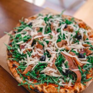Close-up of a delicious arugula and prosciutto pizza with cheese and olives on a wooden table.